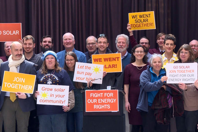 About fifteen people stand holding signs like grow solar in your community and fight for our energy rights