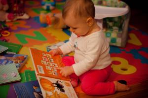 A baby points to a picture book as if reading while sitting on a colorful floor mat 