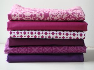 Several bolts of purple fabric in various patterns stacked on top of each other against a white background