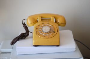 A yellow rotary phone sitting on a stack of white papers against a white wall