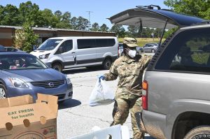 A black woman in army fatigues with face masks loads a full plastic bag into the back of an SUV in a parking lot, with cars lined up in the background.