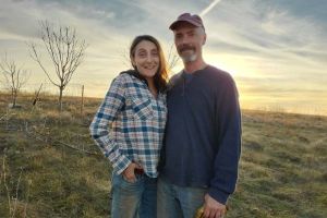 Beth Hoffman and John Hogeland standing close together in a field with a sunset sky, pasture and small trees behind them.