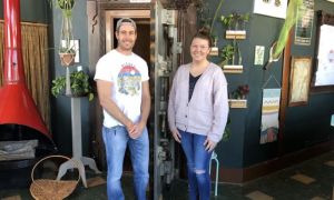 Anthony Gosset and Megan Gosset standing in front of a door to an old bank vault. There are plants and other decor around them including a red metal fireplace.