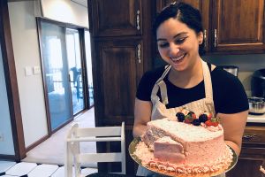 Jennifer Whitley in kitchen with apron holding pink cake decorated with strawberries and blackberrys with a slice taken from the edge of the cake
