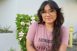 Head shot of Gloria Lucas with long brown hair, large glasses and pink shirt that reads: Eating Disorders are a social justice issue. White background, outdoors, some flowers visible in a shrub