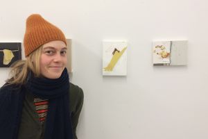 Young woman in winter hat and scarf standing in front of small paintings on a white wall.