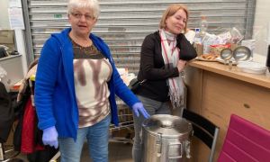 Two women standing in front of a metal garage door, one with her gloved hand over a large metal vat with a clamp lid, the other leaning on a make-shift countertop