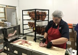 A person in hairnet, apron and gloves handling bacon slices at a meatslicing machine. Cured meat hangs on a rack in the background.