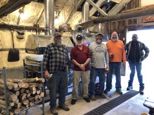 Five white men with beards in ball caps, plaid, and long sleeved t-shirt stand in front of large shiny metal equipment.