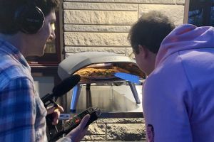 Toby Foster with headphones, a mic and a recorder in hand next to Pete Giordano looking into a small metal oven with a pizza in it. Pete has a knife in his hand. 