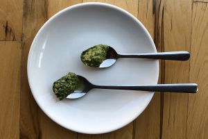 Two metal spoons, one long on shorter, each with a sample of green thick sauce, on a white plate, on a wooden table top in natural lighting