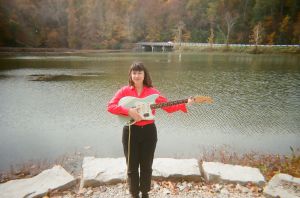 Amy Oelsner stands smiling and holding an electric guitar outside with a lake behind her