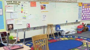 Empty elementary school classroom with a colorful round rug and a whiteboard with writing and posters.