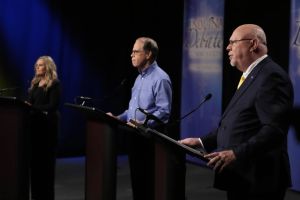 Mike Braun stands on a stage, gesturing while supporters stand behind him. Braun is a White man, balding with gray hair. He is wearing glasses and a blue shirt.