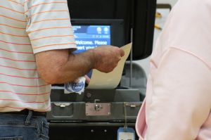 A person feeds a paper ballot into a machine as an election worker is available to assist.