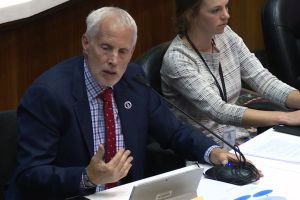 Representative Bob Behning speaks into a microphone during a committee hearing. He wears a navy suit with plaid shirt and red tie. He is a White man with white hair and stubble.