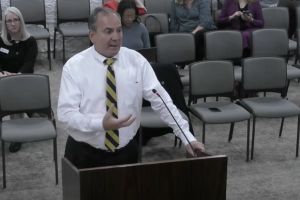 Mike Bohacek stands behind a podium with one hand outstretched. He wears a black and yellow tie with a white button down shirt.