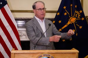 Governor Mike Braun stands at a podium. Behind and flanking him are American and Indiana flags. He wears a grey suit with white shirt. He is a White man, balding, and wears glasses.