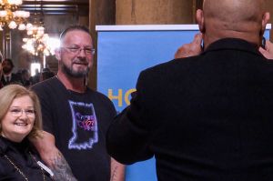 Tony Hostetler is a White man with short hair wearing a Care, Not Cuffs t-shirt. He poses for a picture with attendees at Hoosier Action's event.