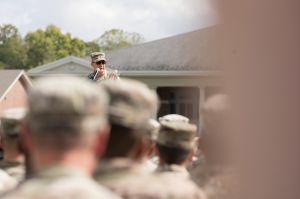 Dale Lyles speaks on a stage in front of a group of National Guard. Lyles is a White man. He is wearing sunglasses and military fatigues.