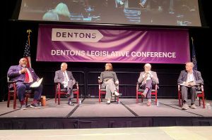 Five people sit on chairs at the front of a stage in front of a purple banner that says "Dentons Legislative Conference."