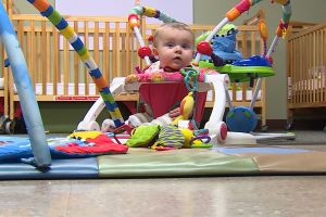 A toddler sits inside a colorful baby bouncer inside a room with cribs in the background.