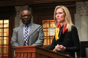 Shelli Yoder speaks into a microphone at a lectern on the Senate floor while Greg Taylor looks on. Yoder is a White woman with blonde hair. She is wearing a black top and a red and yellow scarf. Taylor is a Black man, bald with a goatee.