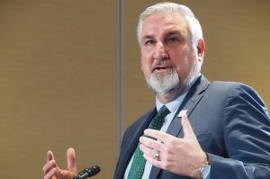 John Keller stands at a podium and speaks into a microphone. Keller is a White man and he is wearing a dark suit with a light blue necktie.