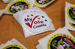 Stickers on a table. The center sticker reads "My Vote Counts" with a large checkmark forming the V.