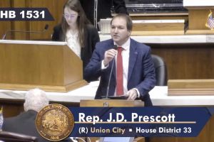 A screenshot of a livestream of the legislature shows J.D. Prescott speaking on the House floor. Prescott is a White man with brown hair. He is wearing a suit and tie.