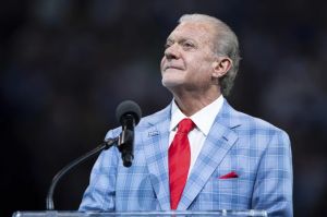 Jim Irsay, a white man with gray hair, speaking at a podium. He's wearing a light blue plaid suit, white shirt and red tie.