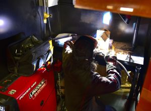 A student with their back to the camera kneels down while practicing welding. To the student's side is the welder, which is red and reads "Lincoln Electric".