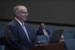 John Keller stands at a podium and speaks into a microphone. Keller is a White man and he is wearing a dark suit with a light blue necktie.