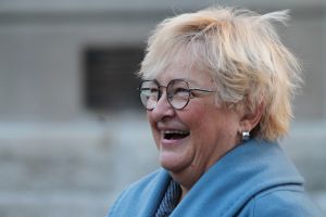 Karen Tallian smiles on the steps of the Indiana Statehouse. Tallian is a White woman with blonde hair. She is wearing glasses and a light blue coat.