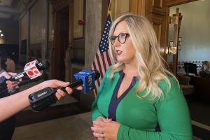 Jennifer McCormick speaks into a bank of microphones during a press conference. McCormick is a White woman with blonde hair. She is wearing a black jacket over a black top.