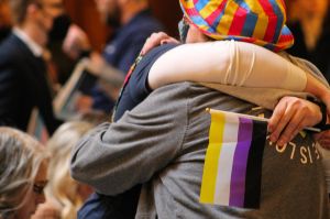 Two people embrace among a crowd inside the Indiana Statehouse. One person has a non-binary pride flag in their hand. The other wears a bucket hat with the pansexual pride colors.