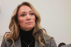 Lindsay Weaver sits in front of a microphone. Weaver is a White woman with dark blonde hair. She is wearing a gray blazer over a black turtleneck.