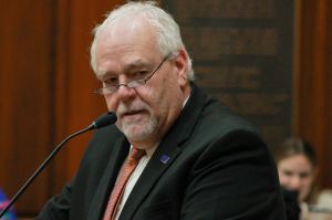 Matt Lehman speaks into a microphone on the floor of the Indiana House. Lehman is a White man with white hair. He is wearing glasses and a black suit.