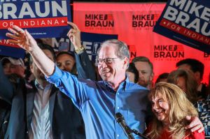 Mike Braun stands on stage at a lectern while a logo for the inauguration ceremony hangs above him. Braun is a White man, balding with gray hair. He is wearing glasses and a suit.