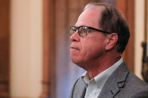 Mike Braun looks upward as he stands in his office. Braun is a White man, balding with gray hair. He is wearing glasses and a gray suit jacket over a white shirt