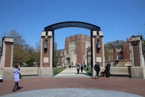 An arch on Purdue's Lafayette campus reads "Purdue University." A brick walkway is in the foreground as people in coats walk through. Behind the arch, a sidewalk connects to other brick buildings.