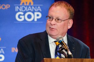 Randy Head speaks into a microphone on a lectern on a stage, with a backdrop of the Indiana GOP logo behind him. Head is a White man with blonde hair. He is wearing glasses and a suit and tie.