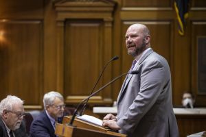 Mike Bohacek stands behind a podium with one hand outstretched. He wears a black and yellow tie with a white button down shirt.