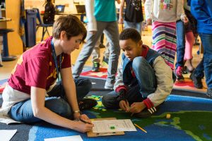 A teacher reviews a student’s completed work in a third-grade classroom as students rotate to new math stations.