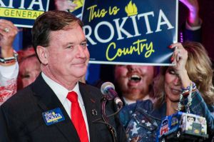 Todd Rokita stands in front of a microphone in front of supporters who are holding signs that read "This is Rokita Country" 