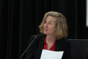 Abby Means stands at a podium holding a packet of papers that have been stapled together. Means is a White woman with shoulder length red hair.